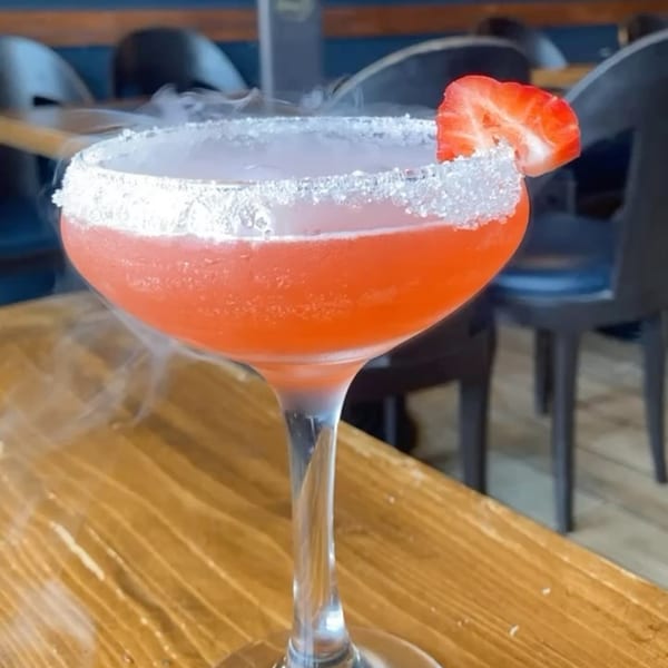 A smoking pink cocktail with a sugared rim and strawberry garnish on a wooden table.