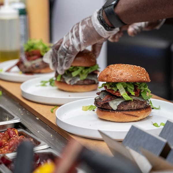 Gloved hands preparing multiple Caribbean-style burgers with cheese, bacon, and lettuce on white plates.