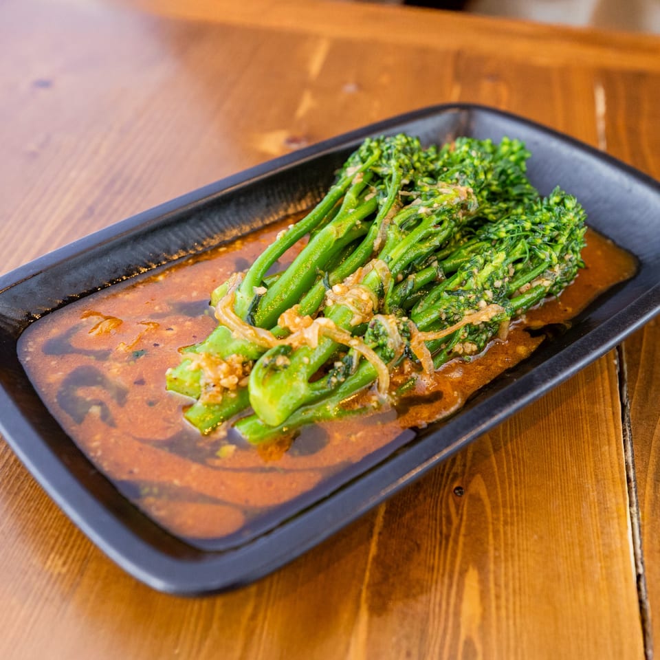 Broccolini with garlic and onions in a rich brown sauce, served on a black rectangular plate.