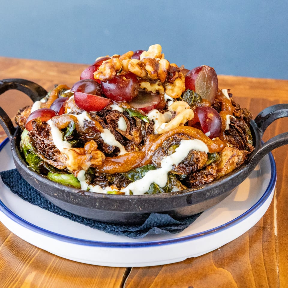 A Caribbean dish of roasted Brussels sprouts, grapes, walnuts, and white sauce in a cast iron pan.