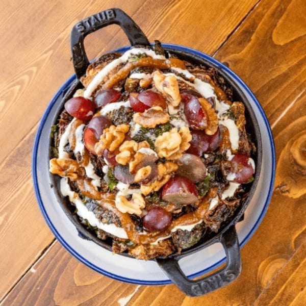 A cast iron skillet of roasted Brussels sprouts, grapes, walnuts, and creamy sauce on a wooden table.